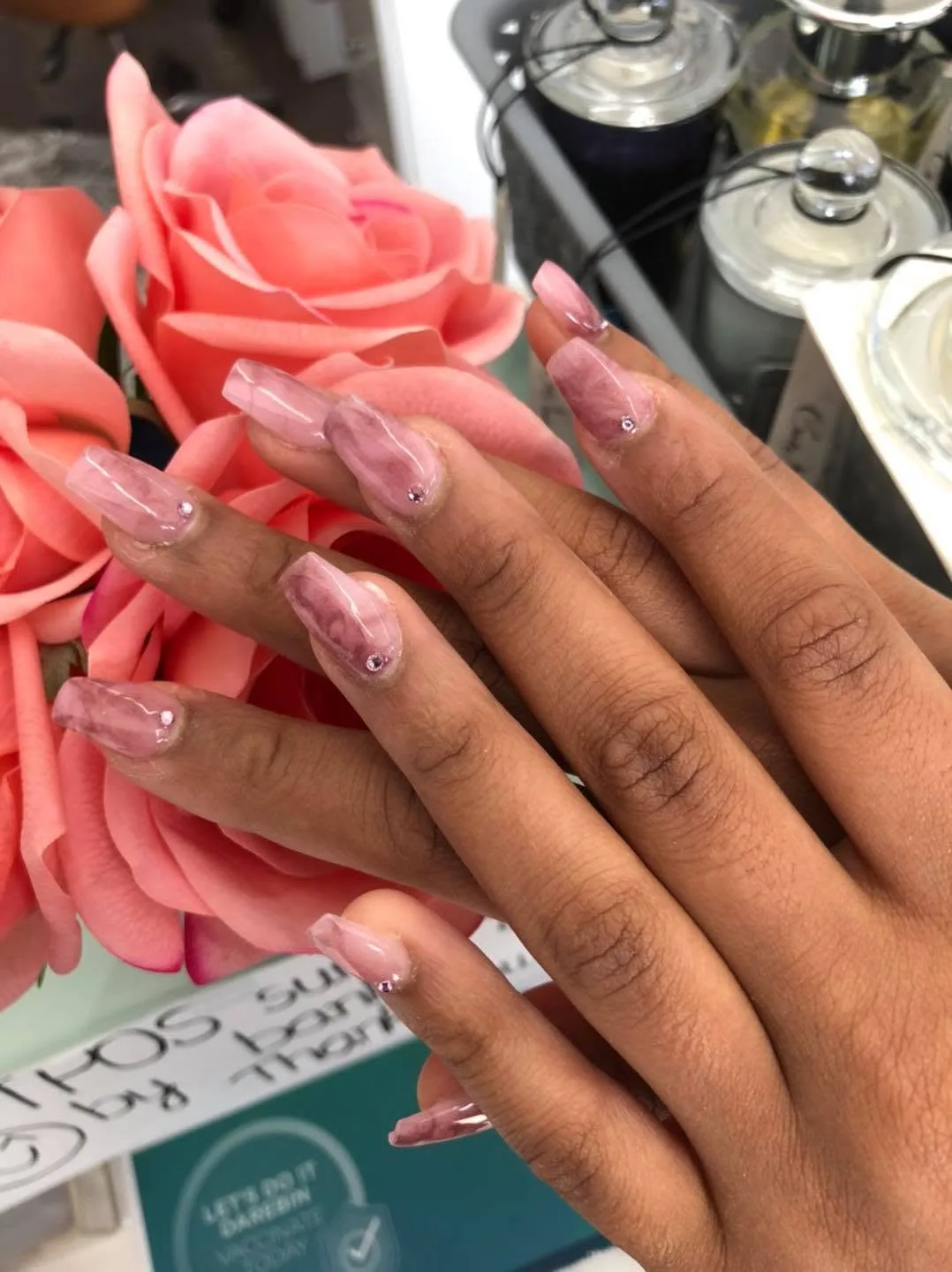 Pink and mauve marble-swirl coffin nails with rhinestones, with coral roses and bokeh bottles behind