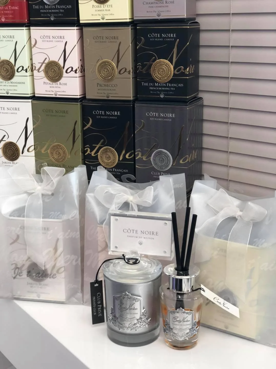 Stacked Côte Noire candle tins with organza gift bags and a reed diffuser by window blinds